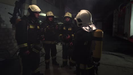 group of five firefighters wearing full equipment in smokey area. relaxed chatting after work well done. fire trucks in the background. name tag translation is fireman and model's last name.