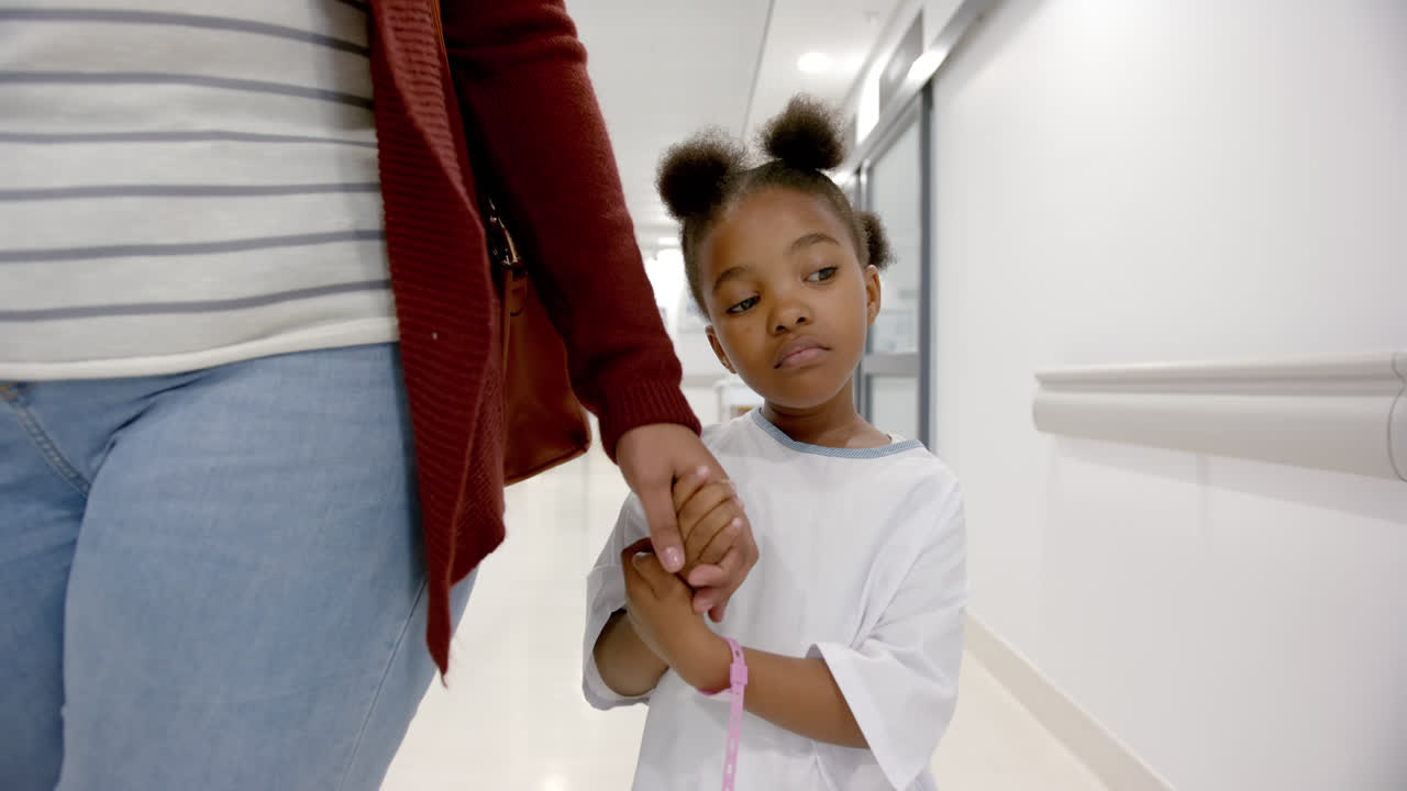 African american mother and daughter in hospital gown holding hands walking in corridor, slow motion