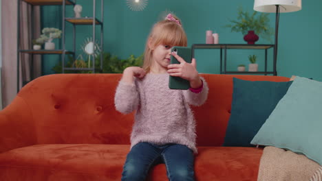 Little-toddler-school-kid-girl-taking-selfie-photo-with-smartphone-while-sitting-on-sofa-at-home