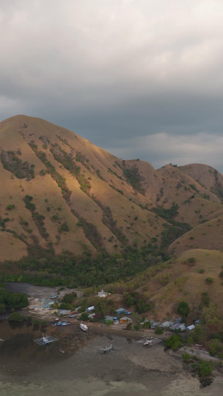 Veduta aerea verticale di un villaggio costiero e di barche da pesca annidati alla base di colline dorate a Komodo