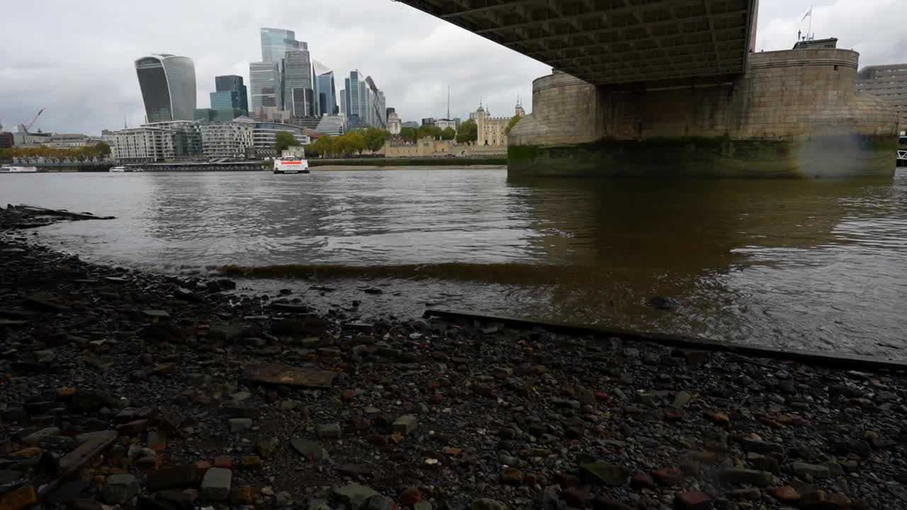 Small waves from passing ships reach the Thames River shore with London’s skyline in the background