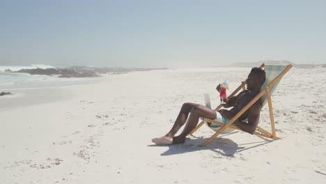 hombre afroamericano usando su computadora portátil en la playa y bebiendo jugo