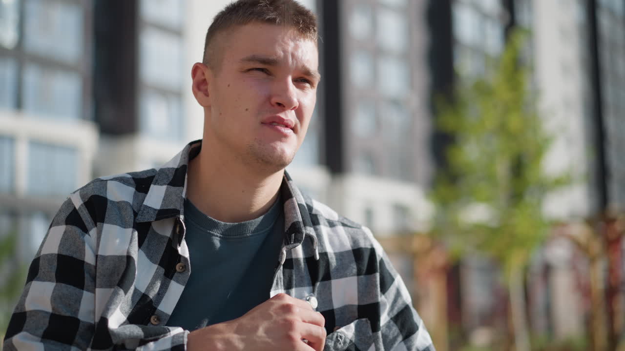 young student in checkered shirt seated outdoors reaches into pocket for medicine with modern tall buildings and blooming flowers in background