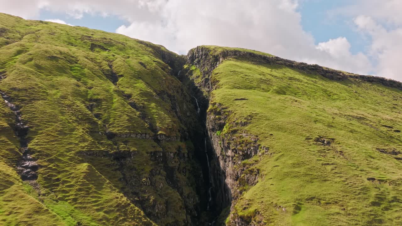 A narrow waterfall flows between green cliffs in the Faroe Islands under soft clouds