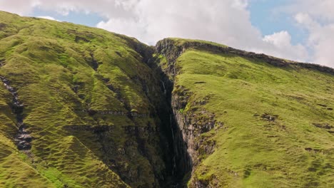 a narrow waterfall flows between green cliffs in the faroe islands under soft clouds