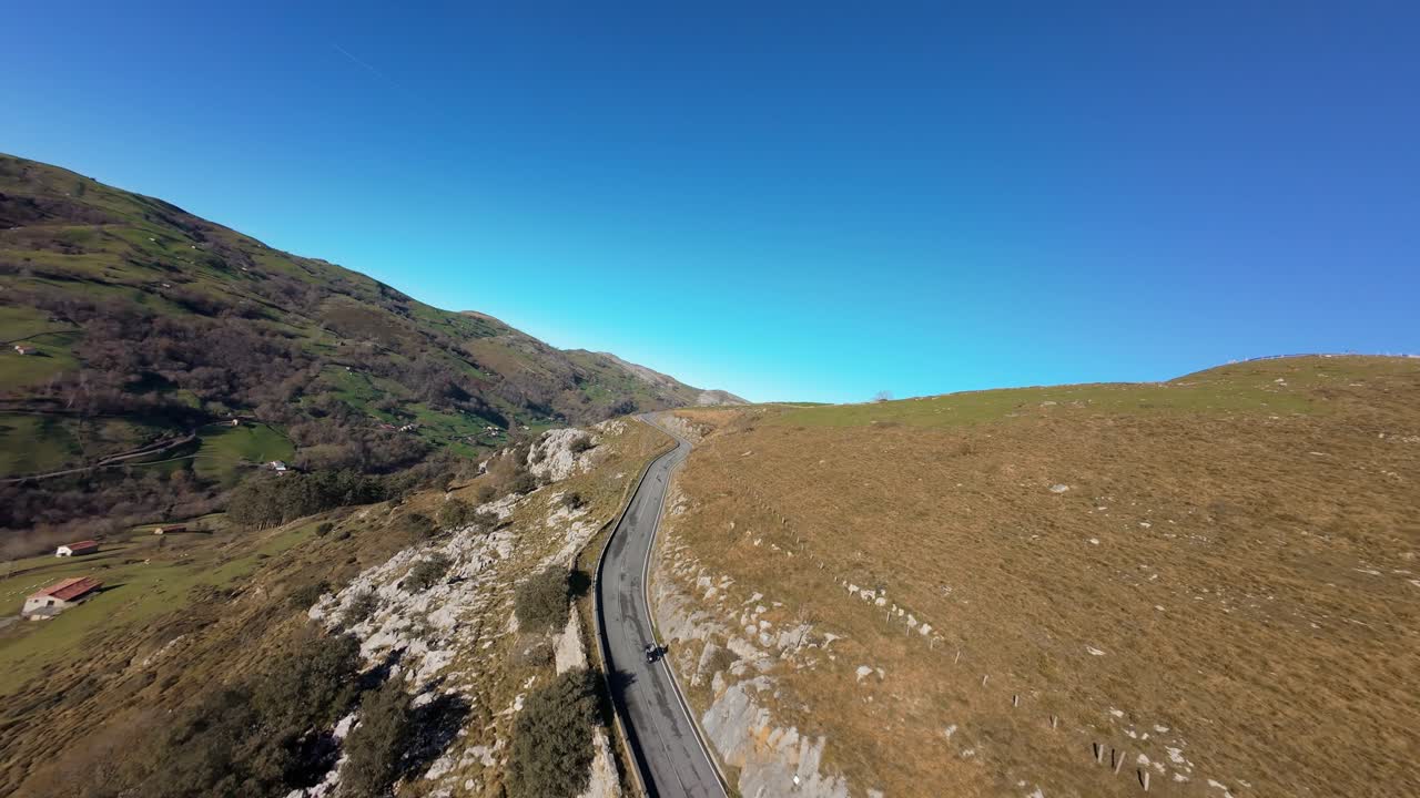 As a motorbike disappears into the distance on a mountain curve, the FPV drone ascends, revealing the vast green valley below.
