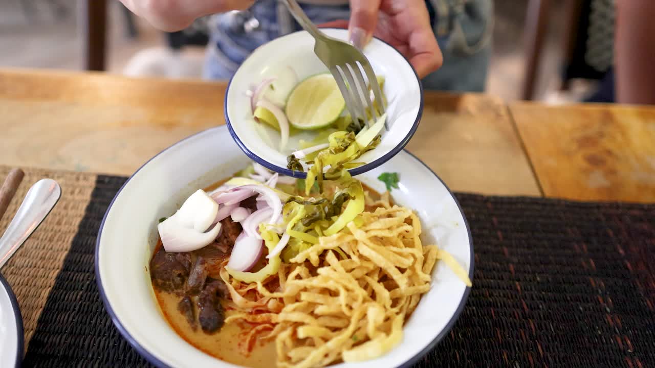 A hand adds pickled vegetables, onions, and lime to a bowl of beef curry noodles in a well-lit Bangkok restaurant, captured in overhead close-up shots