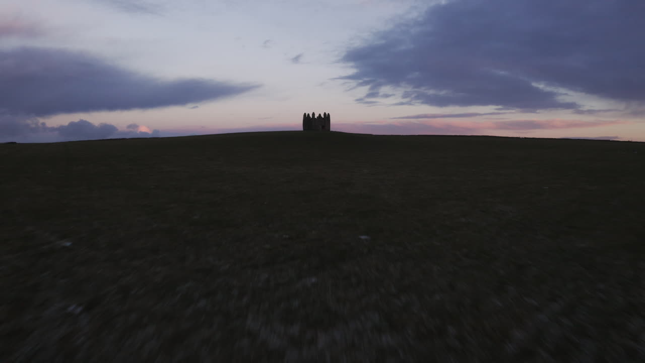Lone castle ruins far in distance under moody twilight sky above darkened open sea, aerial low approach to silhouette