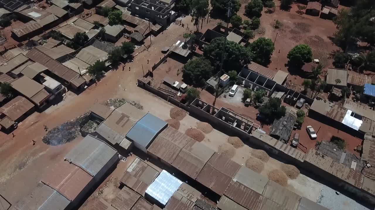 Aerial (drone) view over a neighborhood outside Nampula, Mozambique, showing single-story homes with tin flat roofs, unpaved streets, scattered trees, and a few pedestrians and parked vehicles