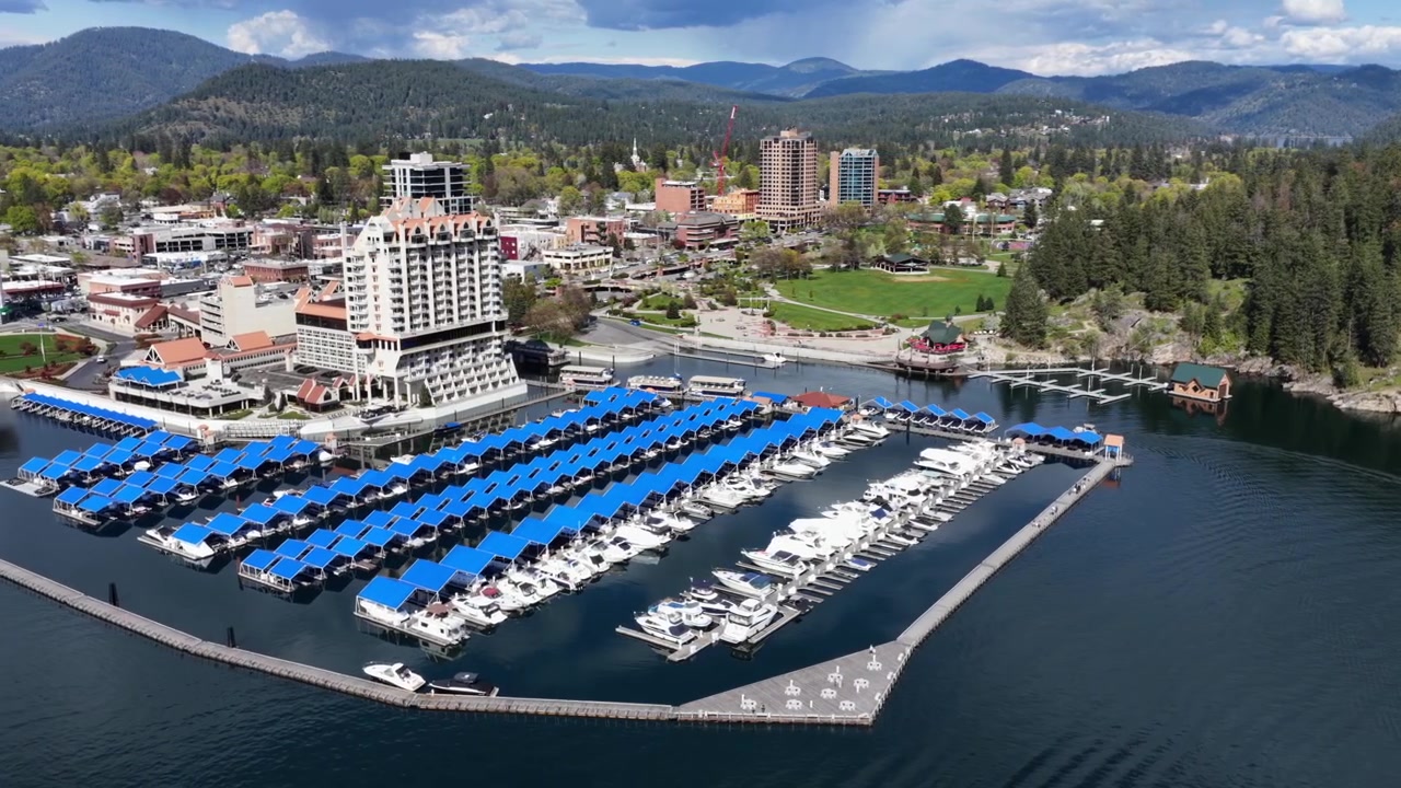 Resort y puerto deportivo de Coeur d'Alene, Idaho, con amarres azules en el lago, edificios del centro y exuberantes pinos verdes bajo un cielo azul brillante en el noroeste del Pacífico de Estados Unidos. Toma aérea orbital amplia.