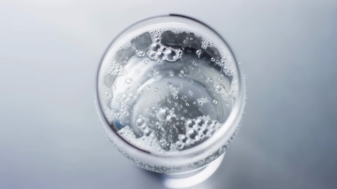 Vista dall'alto di un bicchiere rotondo trasparente che contiene un liquido frizzante e gassato, con bolle dense, una consistenza schiumosa, riflessi luminosi e un'ombra morbida su una superficie grigia minimale per un'atmosfera di bevanda pulita e rinfrescante.