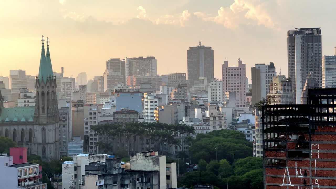 A breathtaking sunset view of the Sé Cathedral in downtown São Paulo. The golden hour light illuminates the cathedral's green copper dome and twin towers against a dense urban skyscraper backdrop
