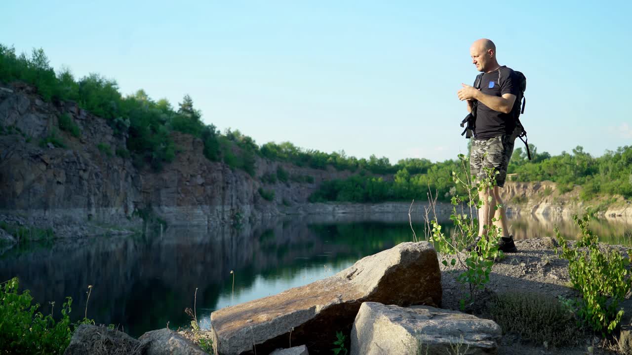 Bald man in t-shirt, shorts and trainers walking with backpack on the big stones near beautiful lake. Natural background of scenic canyon with a clear lake in summer.