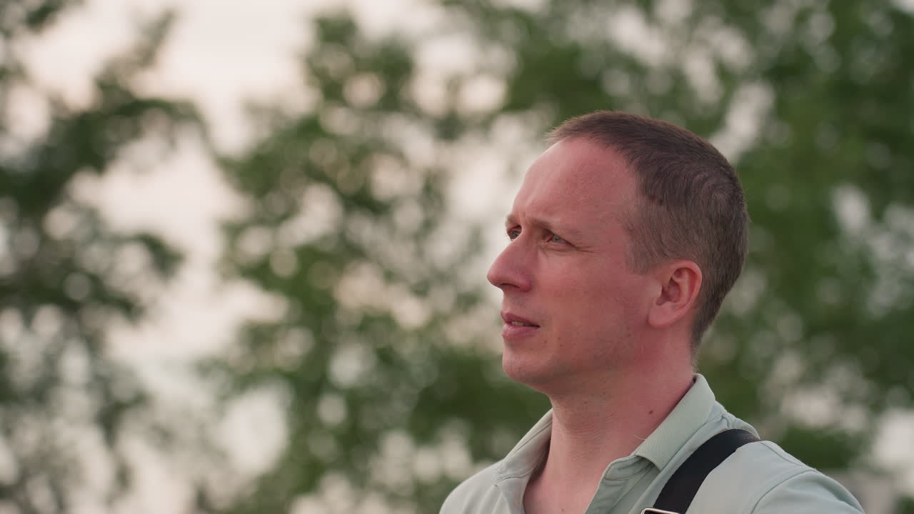 closeup side profile of man with fair skin wearing light grey shirt and shoulder strap looking around thoughtfully against blurred green trees in background