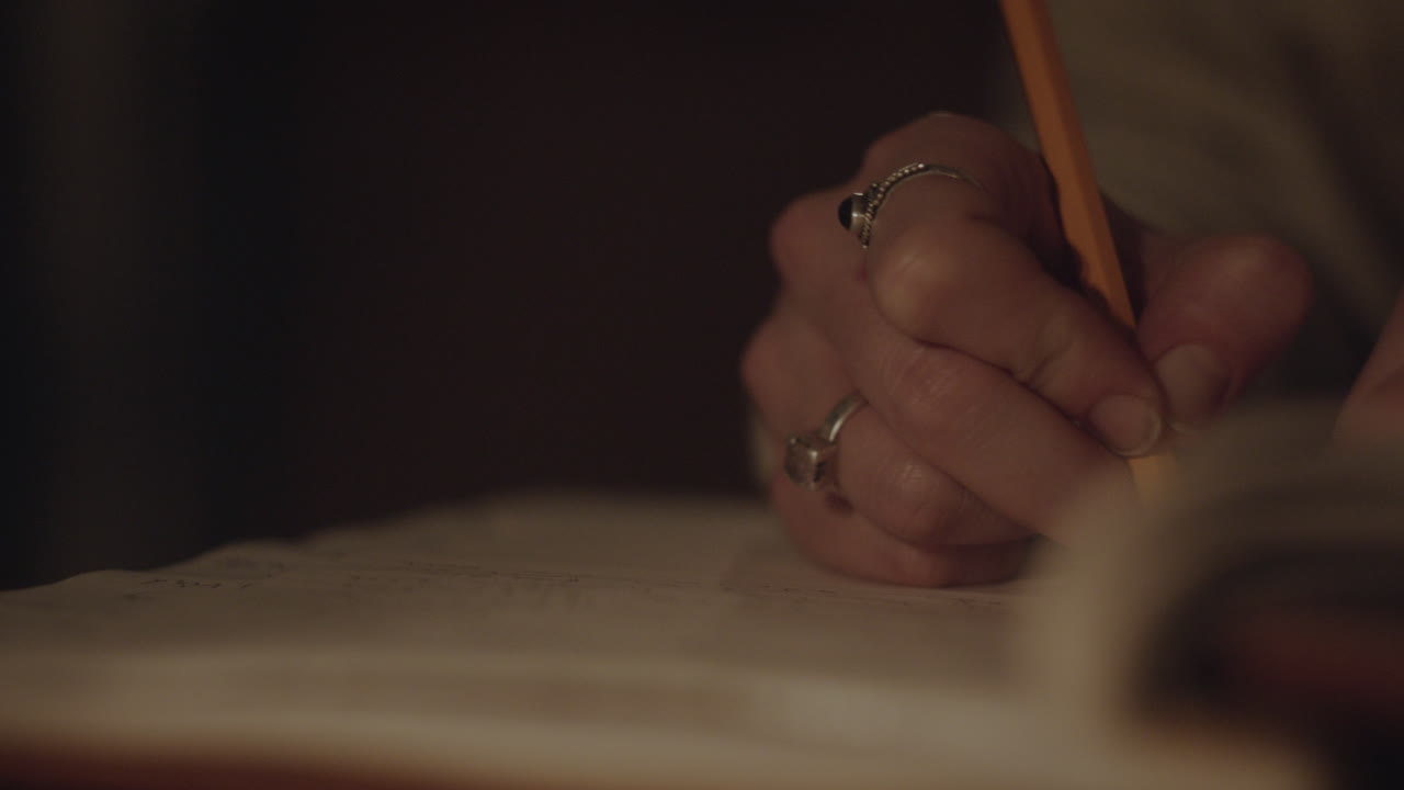 A woman's hand holding a pencil while writing in a journal by candlelight, creating a warm and intimate atmosphere of reflection.