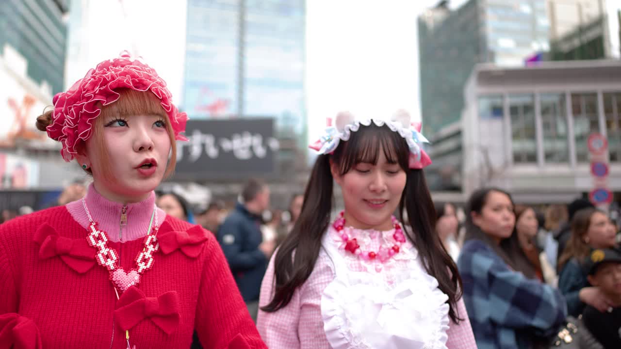 兩位年輕女性穿著色彩繽紛的原宿Decora時尚服飾，在繁忙的澀谷十字路口相視而笑。
