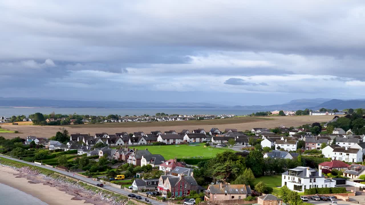 Drone pans above Rosemarkie, revealing beachfront houses, rural scenery, and dramatic cloudy skies