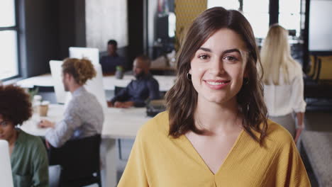 mujer blanca millennial creativa sonriendo a la cámara en una oficina casual ocupada, cabeza y hombros