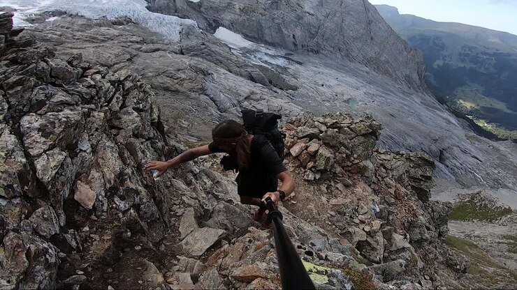 A young, strong and fit man with long hair and tattoos is walking up a steep dirt trail in Switzerland, using both hands and feet to push himself up