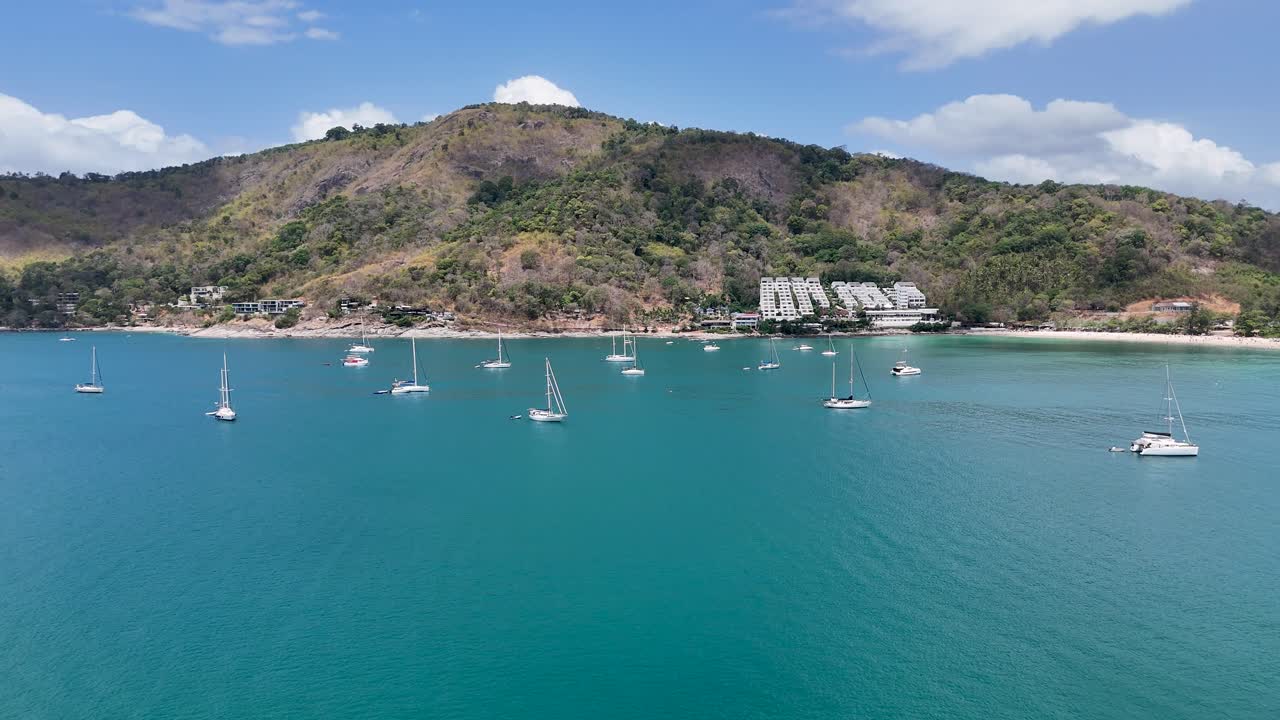 Drone footage captures a serene bay in Phuket, Thailand, showcasing luxury yachts and a picturesque coastal landscape under clear skies