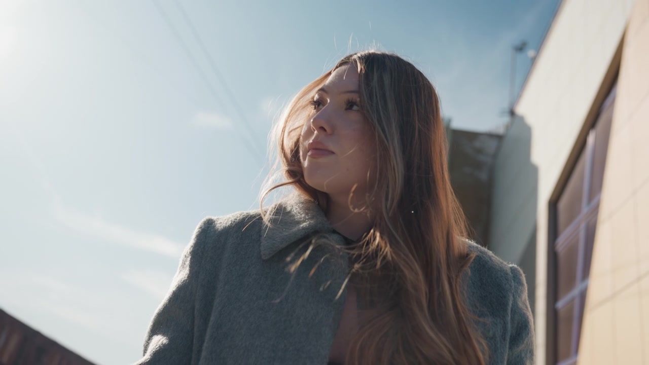 Bonita estudiante asiática dibujando al aire libre, escena urbana soleada en contrapicado con abrigo de lana y cuaderno, mirada pensativa hacia abajo mientras pasa las páginas, contraluz cinematográfico y sombra suave, creatividad delicada.