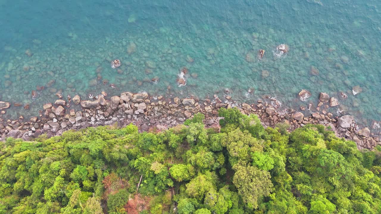 Slow upward drone tilt reveals a rocky coastline with lush green forest meeting the crystal-clear turquoise waters of Koh Rong Sanloem, Cambodia, where a traditional wooden boat floats offshore