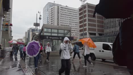 numerous individuals walk along a city street under umbrellas as rain falls, creating a vibrant atmosphere in a bustling urban environment during lunchtime.