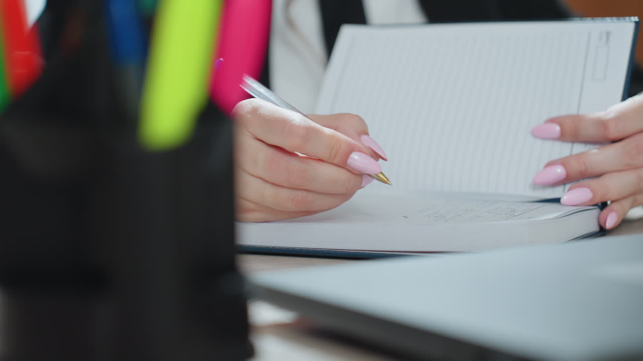 close up of pen container with colorful markers in foreground and partial view of woman with polished pink nails writing in open notebook on office desk with soft natural lighting