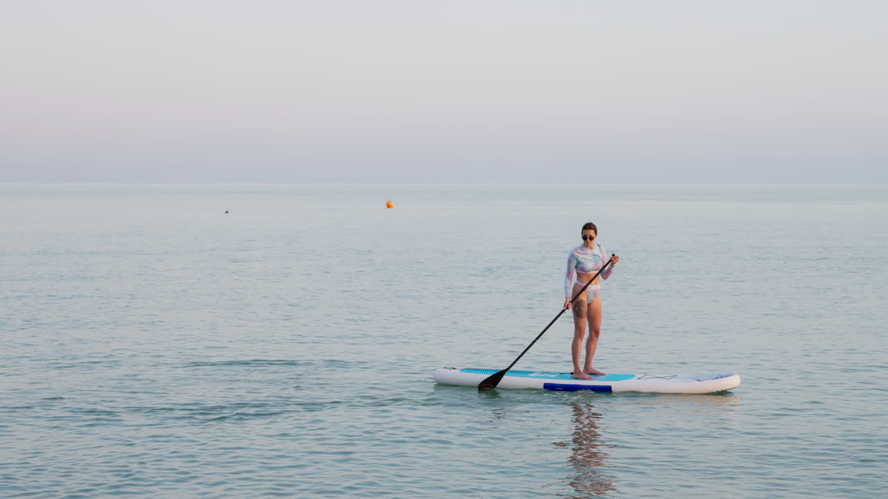 해가 지는 동안 평온한 물에서 패들보드를 타는 여자