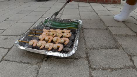 vista bloqueada de la mini parrilla de barbacoa desechable con brochetas de calabacín y camarones mano masculina hurgando y dando la vuelta a la comida