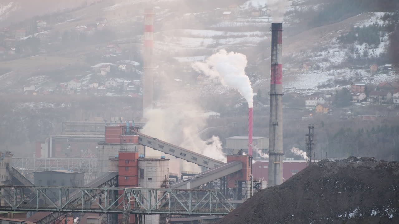 Steam and smoke rise from an industrial complex, with conveyors and a large coal mound in the foreground against a backdrop of a snowy residential hillside