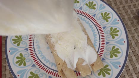 close-up as grated cheese is added over cream-covered mexican taquitos on a decorated plate. static view with warm light.