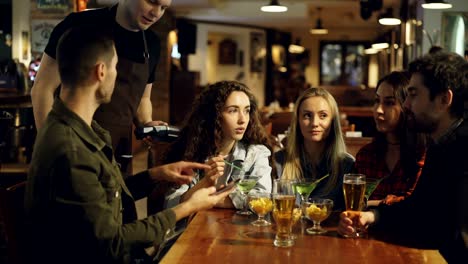 handsome bearded guy is paying with smartphone in pub while having fun with friends. friendly waiter, table with drinks and snacks, nice bar interior are visible.