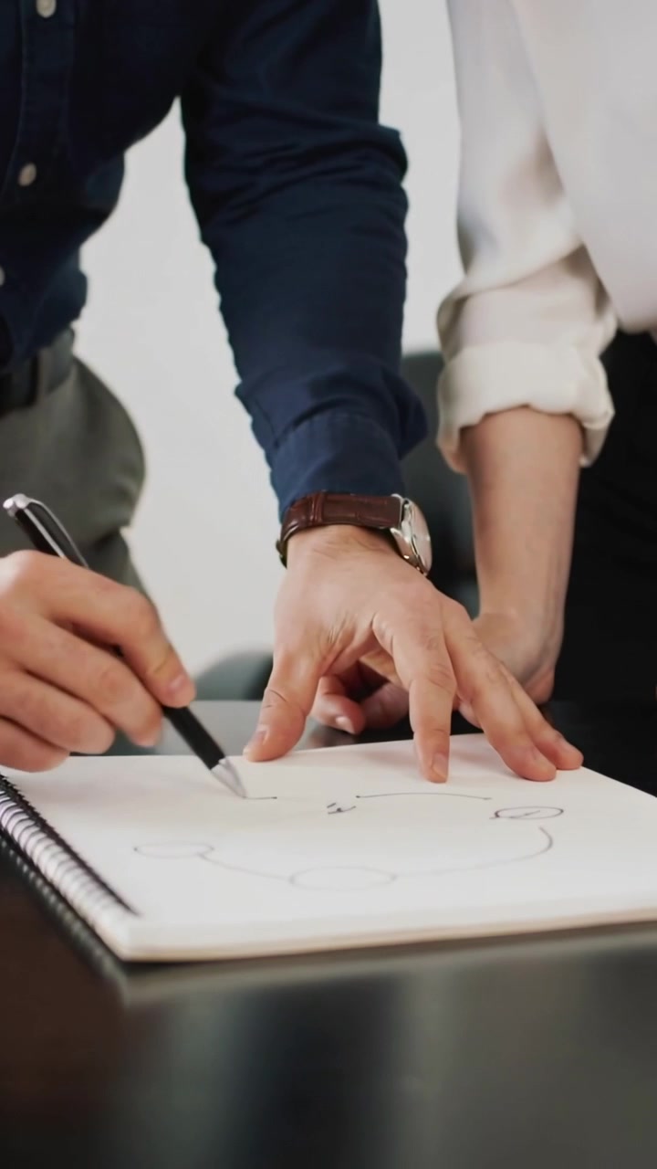 Video verticale: Uomo in camicia appoggia la penna e abbozza un'idea alla scrivania, donna in top indica un blocco a spirale.