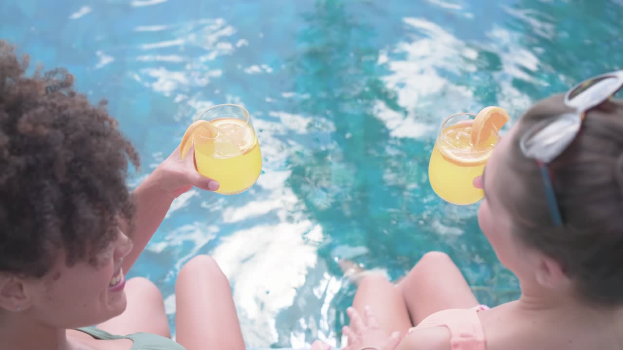 Verschiedene Freundinnen in Badekleidung sitzen am Pool, Kamera zieht sich zurück, stoßen mit gelben Getränken an