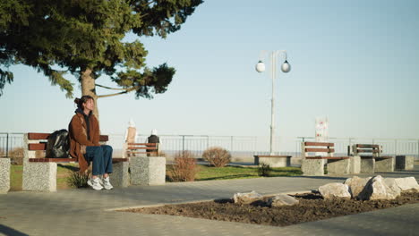 a girl sits alone on a bench in an urban park, looking somber and lost in thought. she is wearing a brown coat, blue jeans, and white shoes, with a black backpack placed beside her