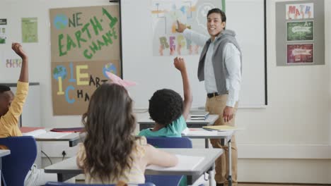 diverse male teacher and children raising hands at desks in elementary school class, slow motion