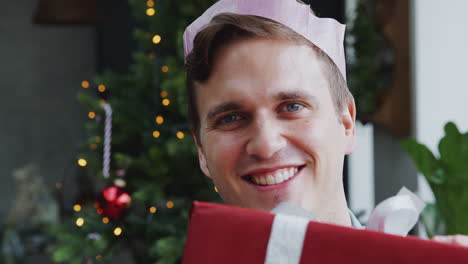 portrait of man wearing paper hat carrying pile of christmas presents standing by tree at home