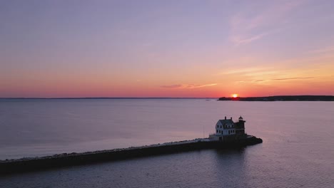 High-aerial-ORBIT-around-a-red-brick-lighthouse-at-the-end-of-a-snow-dusted-rocky-breakwater-with-the-sunrise-and-some-parallax-in-the-distance