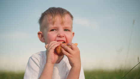 a little boy in a white shirt sits in a grassy field, carefully holding a snack with both hands. he gazes at it intently before taking a bite, enjoying a peaceful moment outdoors in the warm sunshine