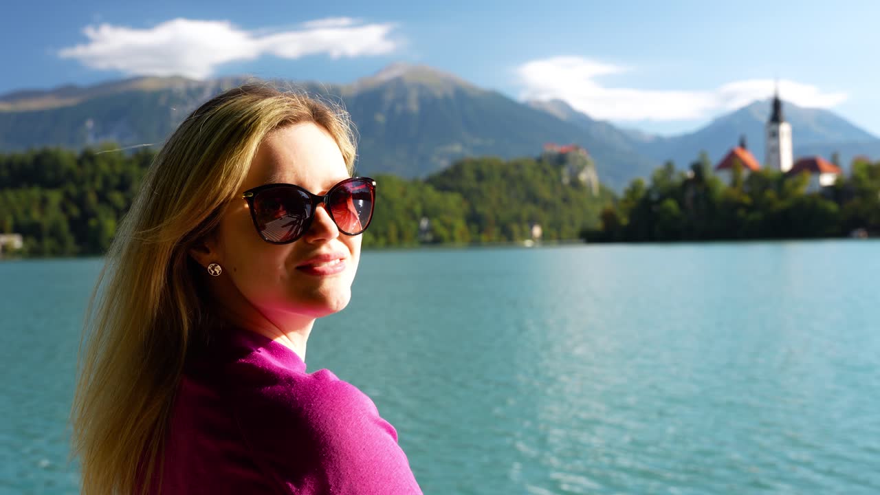 An attractive woman enjoys a peaceful walk beside Lake Bled in Slovenia, with calm water, alpine mountains, and the iconic island church creating a serene travel and lifestyle atmosphere
