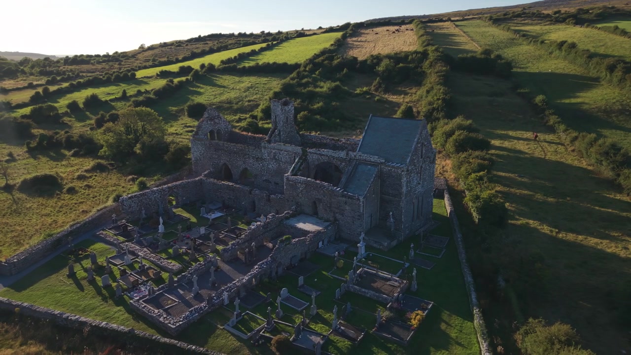 Un'autentica prospettiva aerea delle rovine e del cimitero della storica Abbazia di Corcomroe, circondato da lussureggianti colline verdi irlandesi durante un caldo tramonto.