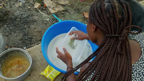 Ghanaian-village-woman-washing-dishes-in-a-soapy-blue-basin-outside-of-her-house-in-Kumasi,-Ghana,-Africa