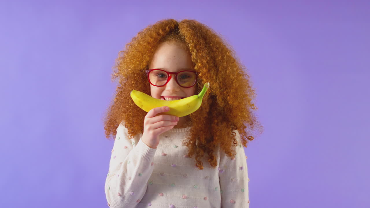 retrato de estudio de una niña sosteniendo un plátano para una boca sonriente contra un fondo púrpura