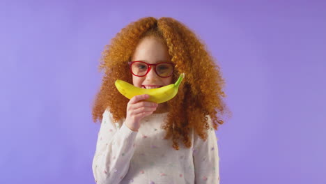 studio portrait of girl holding banana for smiling mouth against purple background