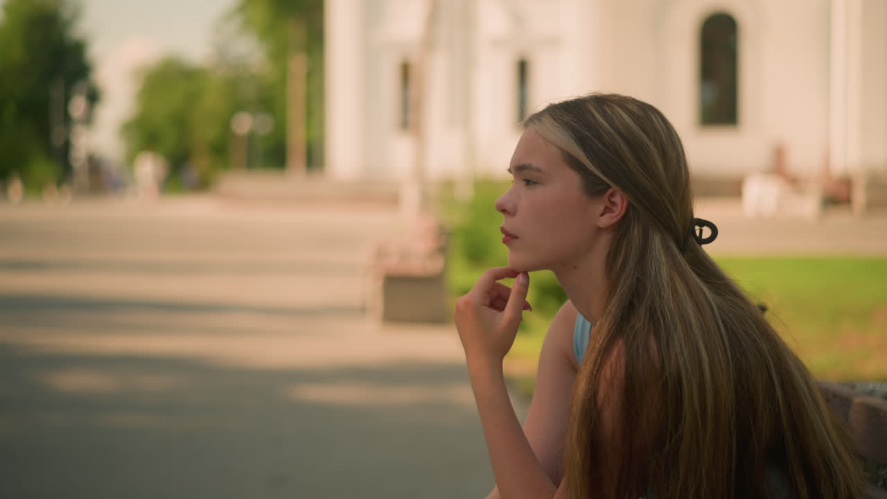 Young lady touching chin with left hand while seated outdoors, gazing thoughtfully into distance, background features building, greenery, and soft shadows under sunlight