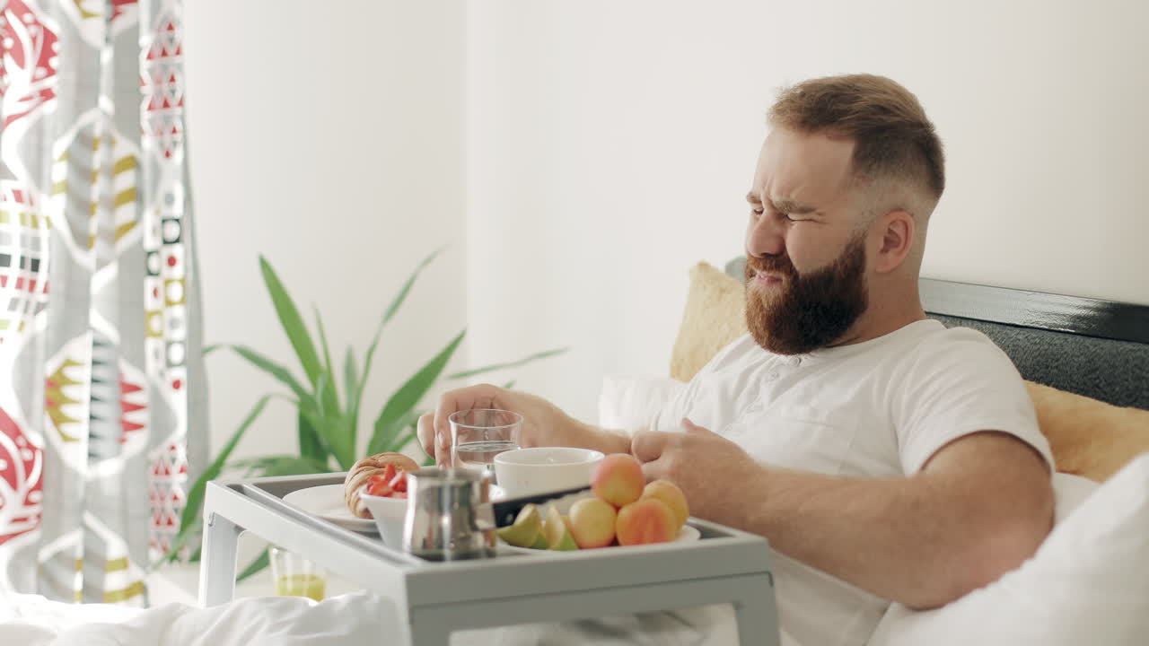 Uomo barbuto che prende pillole e beve acqua mentre fa colazione a letto