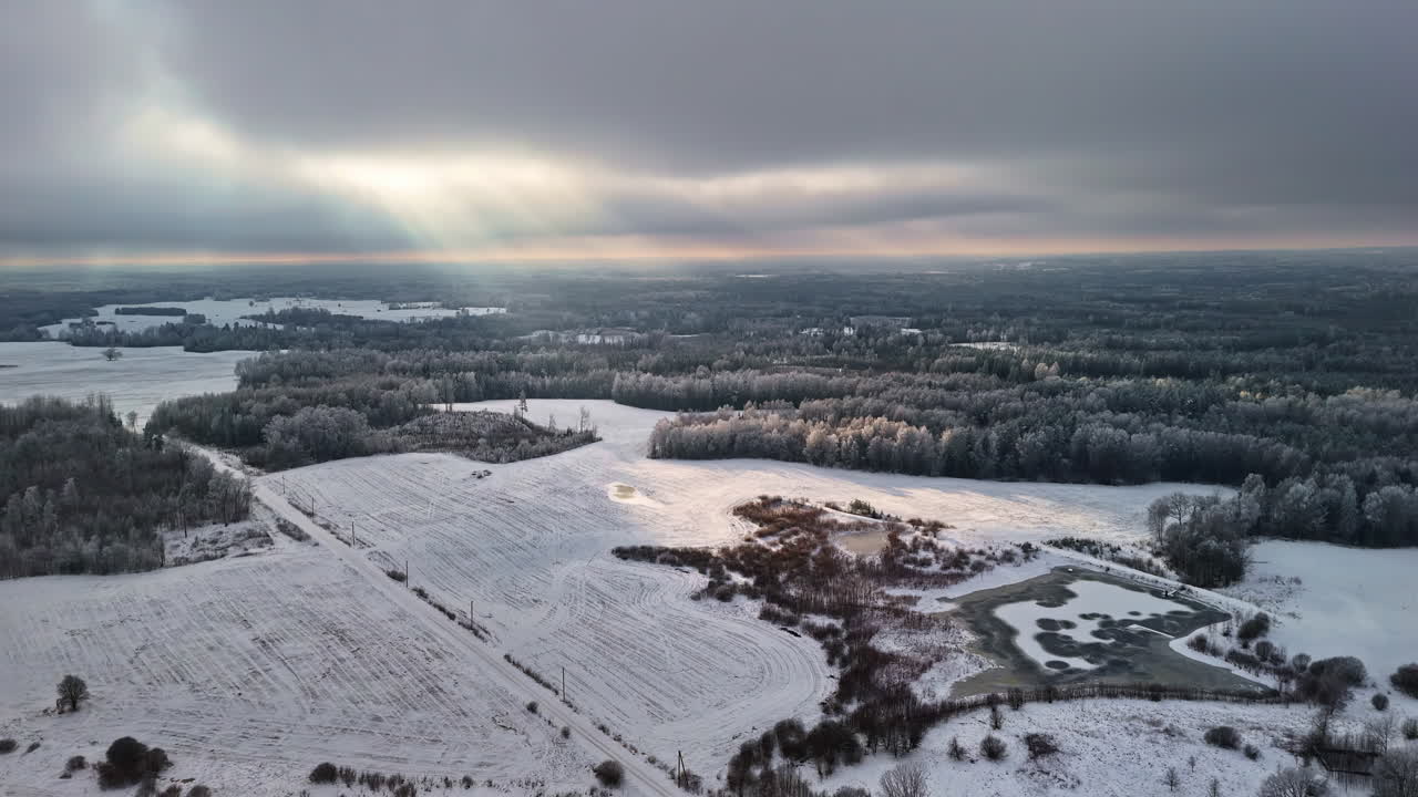 Aerial view of frosty fields, frozen ponds, and dense forest under dramatic winter skies