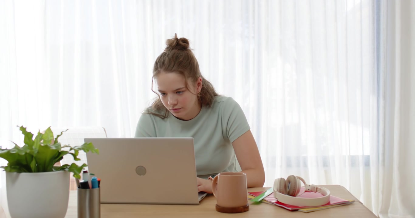 Adult female sitting at desk in mint T-shirt, noticing small text on laptop, leaning forward typing