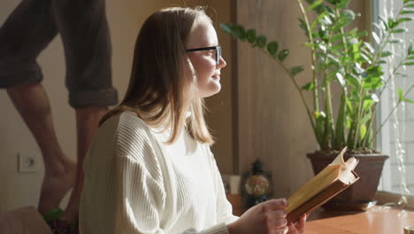 fair skin lady reading book near window bathed in sunlight, blur background shows green potted plant, decorative string lights, mug on table, and wall art